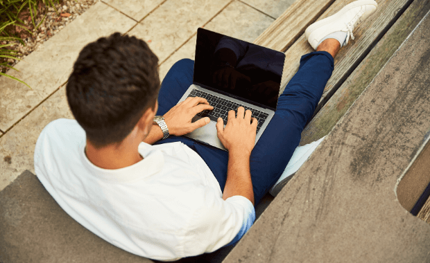 A young man working on his laptop while sitting outside on a park bench.